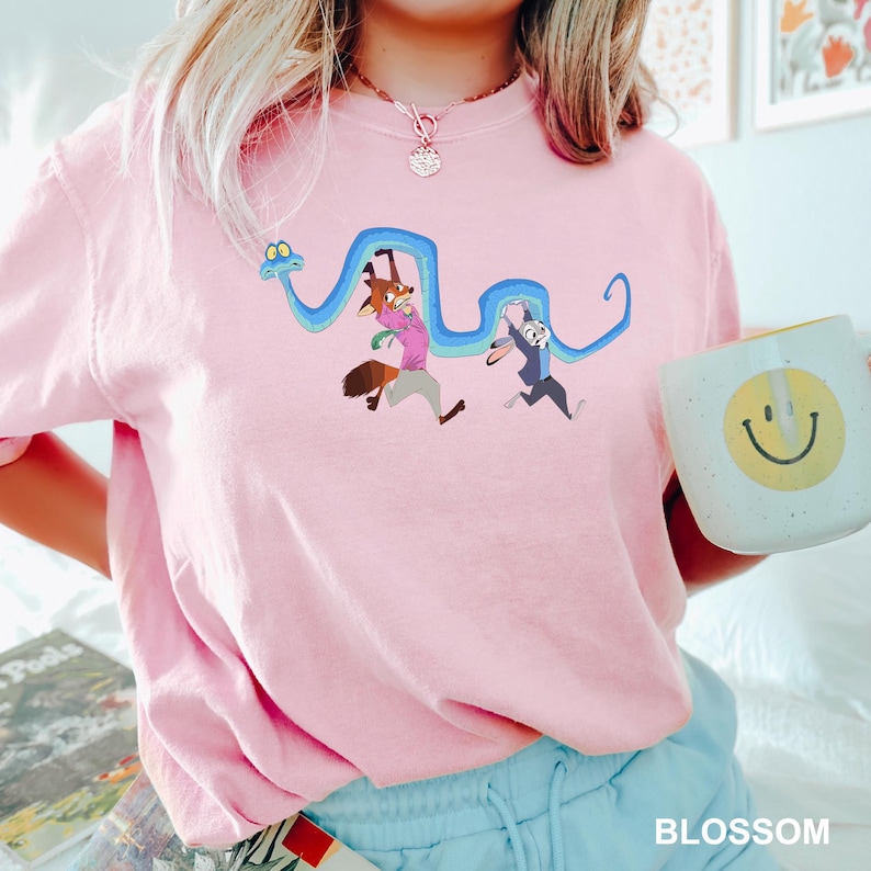 Op de afbeelding: Een lichtroze sweatshirt met een cartoonillustratie van een vos en een konijn die een blauwe slang achtervolgen. De vos draagt een roze shirt en het konijn een blauw pak. Een witte mok met een gele smiley wordt vastgehouden.