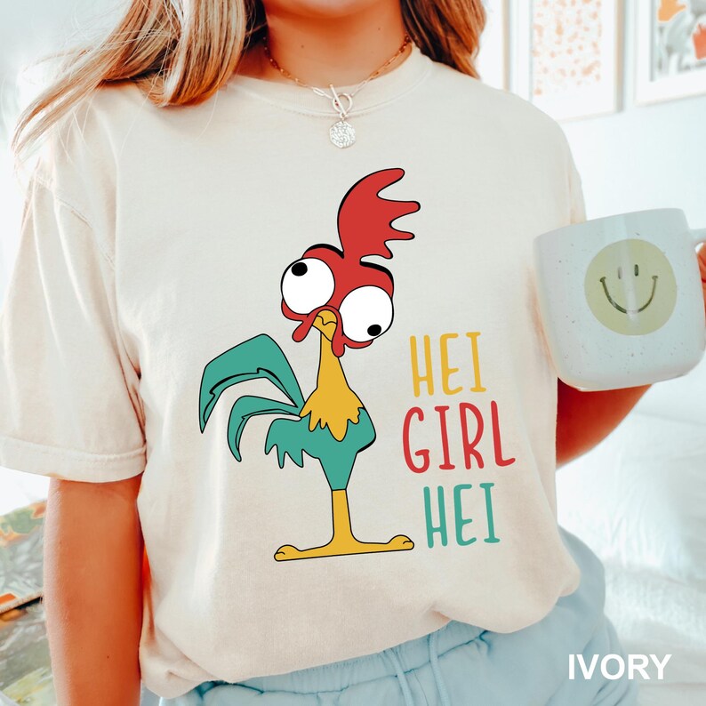 May include: Ivory-colored t-shirt featuring a cartoon rooster with red, yellow, and teal accents. The text "HEI GIRL HEI" is printed in colorful letters. A white mug with a smiley face is also visible.