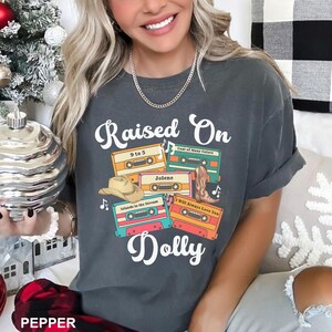 May include: A charcoal gray short-sleeve t-shirt featuring the white text Raised On Dolly. The graphic includes four colorful cassette tapes labeled 9 to 5, Coat of Many Colors, Jolene, Islands in the Stream, and I Will Always Love You.