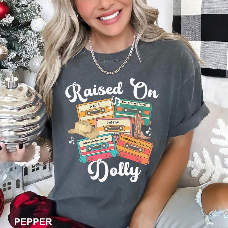 May include: A charcoal gray short-sleeve t-shirt featuring the white text Raised On Dolly. The graphic includes four colorful cassette tapes labeled 9 to 5, Coat of Many Colors, Jolene, Islands in the Stream, and I Will Always Love You.