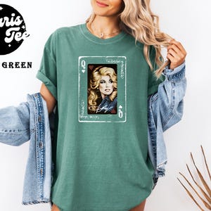 May include: A light green t-shirt featuring a graphic of a playing card with the letter Q in the corners. The card displays a portrait of a blonde person with wavy hair, wearing a denim jacket, with the name Dolly written in white script below.