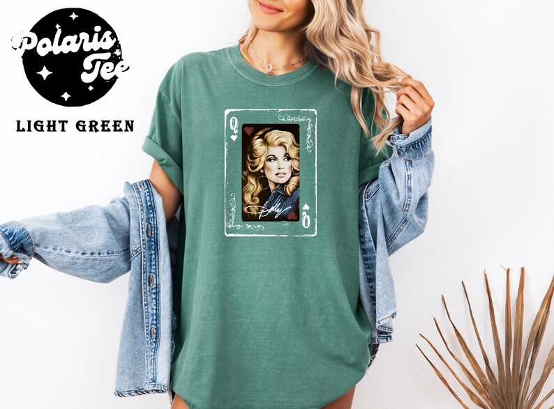 May include: A light green t-shirt featuring a graphic of a playing card with the letter Q in the corners. The card displays a portrait of a blonde person with wavy hair, wearing a denim jacket, with the name Dolly written in white script below.