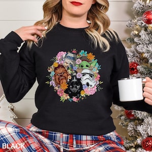 Op de afbeelding: Zwarte crewneck sweatshirt met een bloemenontwerp met Star Wars-personages, waaronder Chewbacca, Darth Vader, een Stormtrooper en Baby Yoda. Het woord "BLACK" staat onderaan gedrukt. Een witte mok wordt in de hand gehouden.