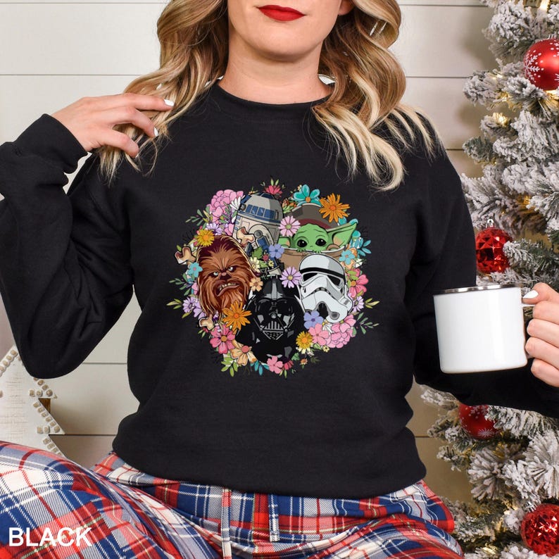 Op de afbeelding: Zwarte crewneck sweatshirt met een bloemenontwerp met Star Wars-personages, waaronder Chewbacca, Darth Vader, een Stormtrooper en Baby Yoda. Het woord "BLACK" staat onderaan gedrukt. Een witte mok wordt in de hand gehouden.