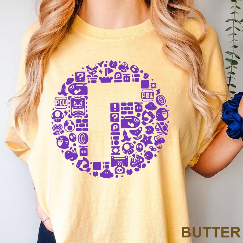May include: A yellow t-shirt with a large purple graphic of the letter "G" filled with various video game icons. The word "BUTTER" is printed in a brown font at the bottom right corner of the shirt.