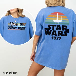 May include: Flo blue t-shirt with a Disney Crew graphic on the front and a Star Wars design on the back. The Star Wars design features a retro sunset, an X-wing, and the text "STAR WARS 1977".