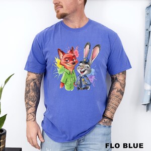 May include: A blue t-shirt featuring a colorful graphic of Nick Wilde and Judy Hopps from Zootopia. Nick is wearing a green shirt, and Judy is in a police uniform. The text "FLO BLUE" is at the bottom of the shirt.