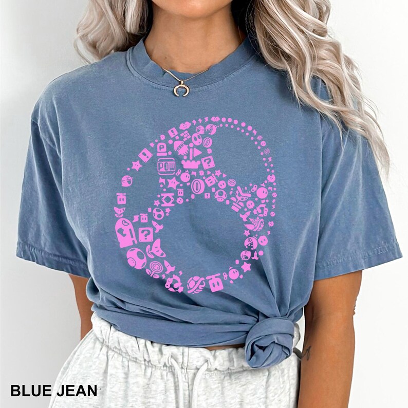 May include: Blue jean-colored t-shirt featuring a pink graphic of a peace sign made up of video game icons. The shirt has short sleeves and a crew neck. The text "BLUE JEAN" is at the bottom of the image.