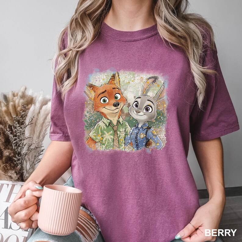 May include: Berry-colored t-shirt featuring a colorful graphic of Nick Wilde and Judy Hopps from Zootopia. Nick is a fox wearing a green shirt, and Judy is a rabbit in a police uniform. The shirt has a relaxed fit.