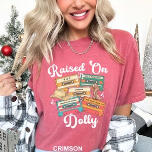 May include: A crimson short-sleeved t-shirt featuring the text Raised On Dolly in white script. The graphic includes four colorful cassette tapes labeled 9 to 5, Coat of Many Colors, Jolene, and I Will Always Love You, with a cowboy hat and boot.