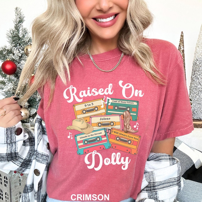 May include: A crimson short-sleeved t-shirt featuring the text Raised On Dolly in white script. The graphic includes four colorful cassette tapes labeled 9 to 5, Coat of Many Colors, Jolene, and I Will Always Love You, with a cowboy hat and boot.