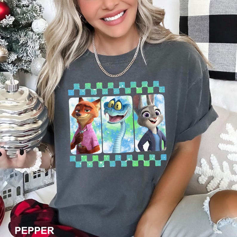 May include: Dark gray t-shirt featuring a graphic of three animated characters: a fox in a pink shirt, a snake, and a rabbit in a blue jacket. The graphic is framed with a green and blue checkered pattern.