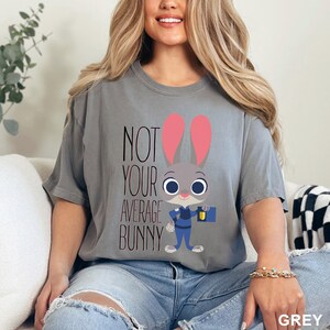 May include: Grey t-shirt with the text "NOT YOUR AVERAGE BUNNY" and a cartoon bunny character wearing a police uniform. The bunny has pink ears and is holding a briefcase and a flashlight.