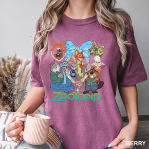 K&ouml;nnte beinhalten: Beerenfarbenes T-Shirt mit einer farbenfrohen Grafik von Zootopia-Charakteren in einem dekorativen Rahmen. Das Wort "ZOOTOPIA" ist darunter in Gr&uuml;n und Blau gedruckt. Das Shirt ist ein bequemer, l&auml;ssiger Stil.