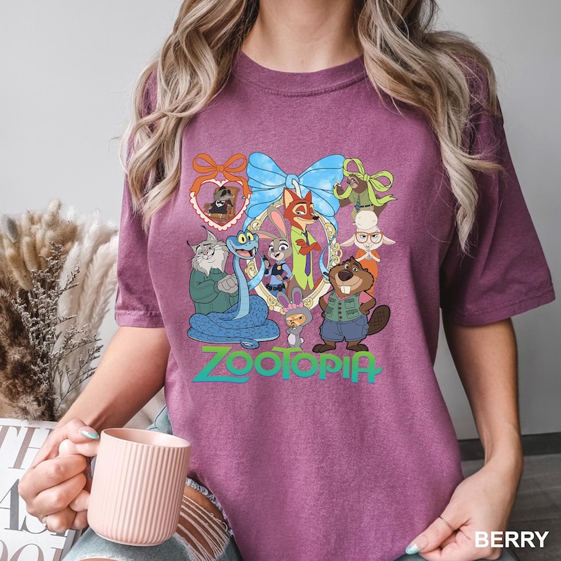 K&ouml;nnte beinhalten: Beerenfarbenes T-Shirt mit einer farbenfrohen Grafik von Zootopia-Charakteren in einem dekorativen Rahmen. Das Wort "ZOOTOPIA" ist darunter in Gr&uuml;n und Blau gedruckt. Das Shirt ist ein bequemer, l&auml;ssiger Stil.