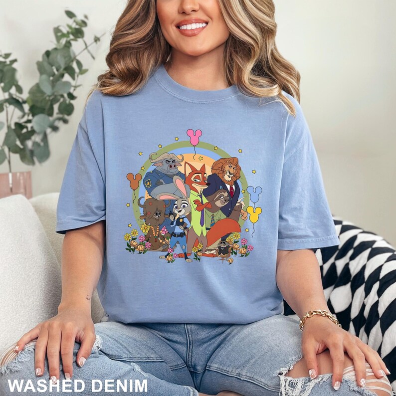 K&ouml;nnte beinhalten: Hellblaues T-Shirt mit einem farbenfrohen Cartoon-Motiv von Zootopia-Charakteren, darunter Judy Hopps und Nick Wilde. Die Grafik ist von einem Regenbogen und Blumen umgeben. Der Text "WASHED DENIM" befindet sich am unteren Bildrand.