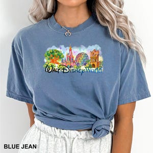 May include: A blue jean colored short-sleeve t-shirt featuring a colorful graphic print of the Walt Disney World theme park landmarks, including a castle, a geodesic sphere, and a tree, with the text Walt Disney World printed in blue.