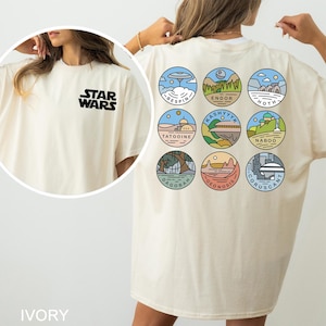 May include: Ivory-colored t-shirt featuring the Star Wars logo on the front. The back displays nine circular illustrations of planets from the Star Wars universe, each with a unique landscape and name. The shirt is a relaxed fit.