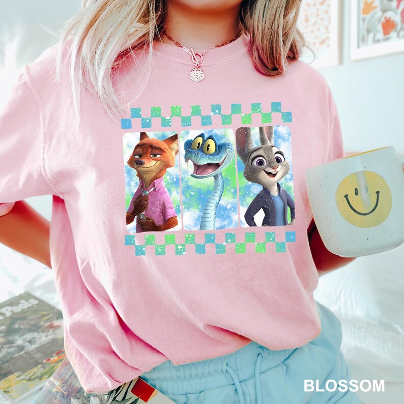 May include: A light pink t-shirt featuring three cartoon characters in a checkered frame. The characters are a fox, a snake, and a rabbit. A white mug with a yellow smiley face is on the right side of the image. The word "BLOSSOM" is at the bottom.