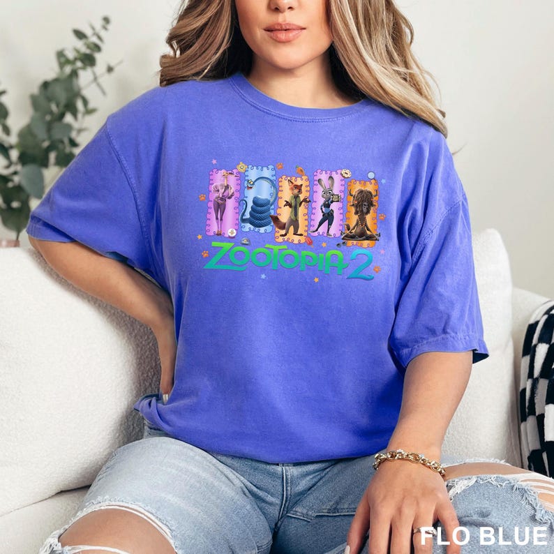 K&ouml;nnte beinhalten: Ein blaues T-Shirt mit einer farbenfrohen Grafik aus dem Film Zootopia 2. Die Grafik zeigt mehrere Charaktere in rechteckigen Rahmen, mit dem Filmtitel in Gr&uuml;n. Das Shirt hat eine einfarbige blaue Farbe.