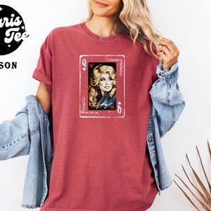 May include: A crimson short-sleeve t-shirt featuring a graphic print of a playing card with a queen of hearts design. The center of the card displays a portrait of a blonde woman with a signature below it, framed by a white border on the shirt.
