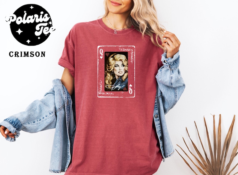 May include: A crimson short-sleeve t-shirt featuring a graphic print of a playing card with a queen of hearts design. The center of the card displays a portrait of a blonde woman with a signature below it, framed by a white border on the shirt.