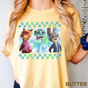 May include: Yellow t-shirt featuring a graphic of three animated characters: a fox in a pink shirt, a blue snake, and a rabbit in a blue jacket. The design is framed by a checkered border.