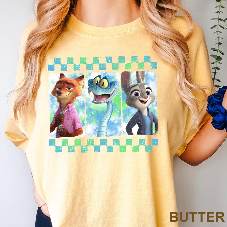 May include: Yellow t-shirt featuring a graphic of three animated characters: a fox in a pink shirt, a blue snake, and a rabbit in a blue jacket. The design is framed by a checkered border.