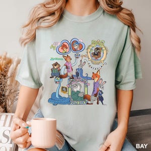 May include: A pale green t-shirt with a vibrant graphic of Zootopia characters. The design showcases animal characters within decorative frames, along with the word "Zootopia". The t-shirt has a crew neck.