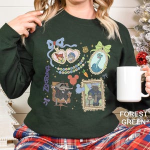 以下が含まれることがあります： ズートピアのキャラクターの額入りイラストが特徴のフォレストグリーンのスウェットシャツ。デザインには、ニック・ワイルドとジュディ・ホップス、ヘビ、その他のキャラクターが含まれています。「ズートピア」の文字がスウェットシャツの左側に縦にプリントされています。