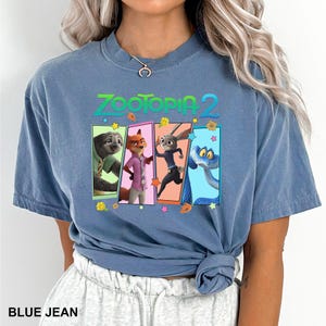 May include: Blue jean colored t-shirt featuring the text "ZOOTOPIA 2" in green and orange. The shirt displays four character panels from the movie. The bottom of the shirt is knotted.