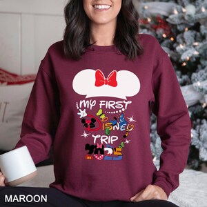 Puede incluir: Sudadera granate con la frase "My First Disney Trip 2025". El dise&ntilde;o incluye orejas de Mickey Mouse con un lazo rojo y varias ilustraciones de personajes de Disney en colores brillantes. La palabra "MAROON" est&aacute; en la parte inferior.