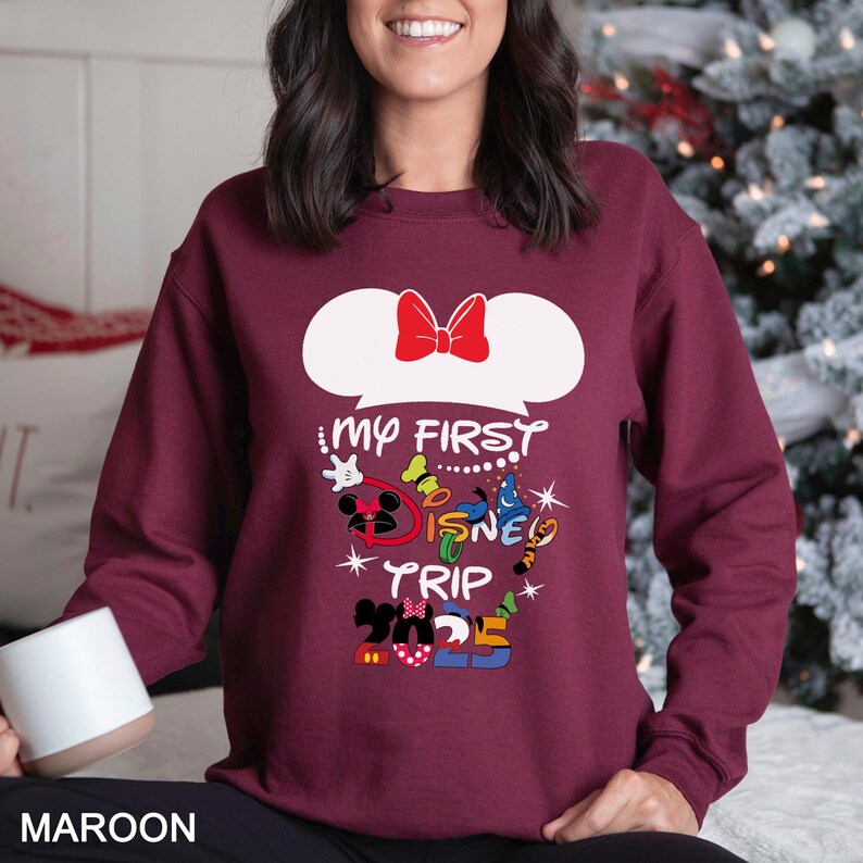 Puede incluir: Sudadera granate con la frase "My First Disney Trip 2025". El dise&ntilde;o incluye orejas de Mickey Mouse con un lazo rojo y varias ilustraciones de personajes de Disney en colores brillantes. La palabra "MAROON" est&aacute; en la parte inferior.