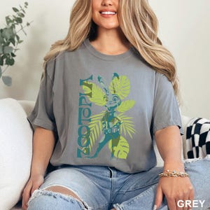 May include: A grey t-shirt featuring a cartoon character from Zootopia. The design includes the word "Zootopia" in teal, along with a cartoon image of a rabbit character and tropical leaves in shades of green. The shirt is a relaxed fit.