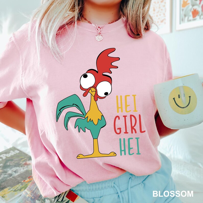 May include: A light pink t-shirt featuring a cartoon rooster with red, yellow, and teal accents. The text "HEI GIRL HEI" is printed in colorful letters. A white mug with a yellow smiley face is held in the image.