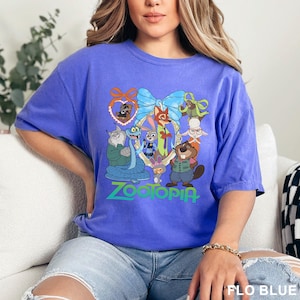 K&ouml;nnte beinhalten: Ein blaues T-Shirt mit einer farbenfrohen Grafik von Charakteren aus dem Animationsfilm Zootopia. Das Design umfasst Judy Hopps, Nick Wilde und andere Charaktere. Das Wort "Zootopia" ist unten gr&uuml;n gedruckt.