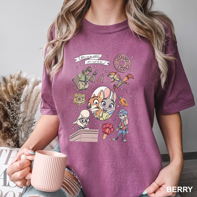 Op de afbeelding: Bessenkleurig T-shirt met een kleurrijke afbeelding van personages uit een populaire animatiefilm. Het ontwerp bevat een donut, een politiebadge en verschillende dierenfiguren. De tekst op het shirt luidt "You can be whatever you wish to be."
