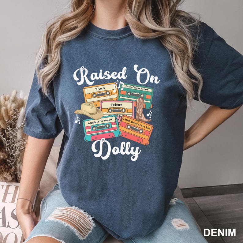 May include: A dark blue t-shirt featuring a graphic print of four colorful cassette tapes labeled with song titles including 9 to 5, Jolene, Islands in the Stream, and I Will Always Love You, along with a cowboy hat, a boot, and the text Raised On Dolly.