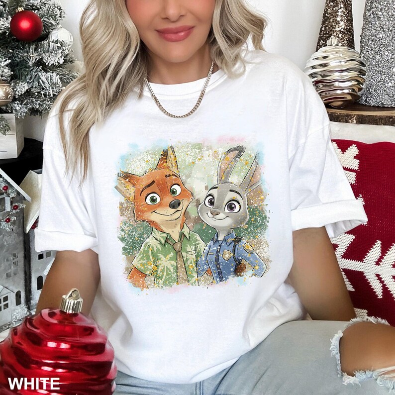 May include: White t-shirt featuring a colorful graphic of Nick Wilde and Judy Hopps from Zootopia. Nick is a fox wearing a green shirt, and Judy is a rabbit in a police uniform. The shirt has a relaxed fit.