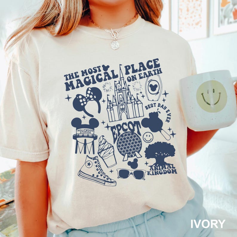 Puede incluir: Camiseta color marfil con un gr&aacute;fico azul marino que presenta iconos de Disney y texto: "The Most Magical Place on Earth", "Epcot" y "Animal Kingdom". Se sostiene una taza blanca con una carita sonriente.
