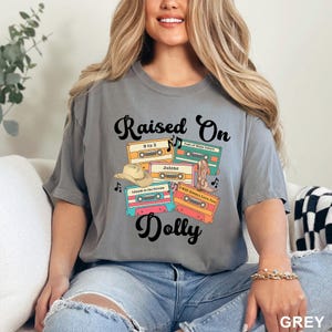 May include: A grey short-sleeve t-shirt featuring a graphic print with the text "Raised On Dolly" and four colorful cassette tapes labeled "9 to 5", "Coat of Many Colors", "Jolene", and "I Will Always Love You", plus a cowboy hat and boot.