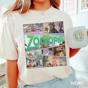 K&ouml;nnte beinhalten: Ein cremefarbenes Kurzarm-T-Shirt mit einem mehrteiligen Grafikdruck aus dem Film Zoomania. Das Design zeigt verschiedene Szenen mit den Charakteren Nick Wilde und Judy Hopps sowie den gro&szlig;en gr&uuml;nen Zoomania-Filmlogo-Schriftzug.