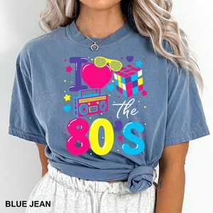 K&ouml;nnte beinhalten: Blaues Jeans-T-Shirt mit einem farbenfrohen Grafikdesign mit der Aufschrift "I love the 80s". Das Design umfasst ein rosa Herz, gelbe Sonnenbrillen, einen Rubik's Cube und einen Ghettoblaster. Der Text "BLUE JEAN" befindet sich unten.