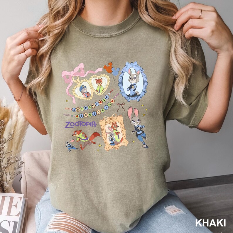 Op de afbeelding: Khaki T-shirt met kleurrijke cartoon illustraties uit de film "Zootopia". Het ontwerp bevat ingelijste karakterportretten, een roze strik en het woord "Zootopia" in een speels lettertype. Het shirt is een comfortabele, casual stijl.
