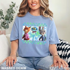 May include: A light blue t-shirt featuring three cartoon characters: a fox in a pink shirt, a snake, and a rabbit in a blue jacket. The shirt has the words "WASHED DENIM" at the bottom.