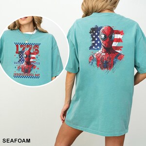 May include: A teal t-shirt featuring a graphic of a red and blue superhero mask over a stylized American flag. The back displays the large graphic, while the front inset shows a smaller version with the text 1776 UNITED STATES INDEPENDENCE DAY.