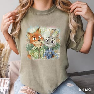 May include: Khaki-colored t-shirt featuring a colorful graphic of Nick Wilde and Judy Hopps from Zootopia. Nick is wearing a green shirt, and Judy is in a police uniform. The word "KHAKI" is printed at the bottom right.