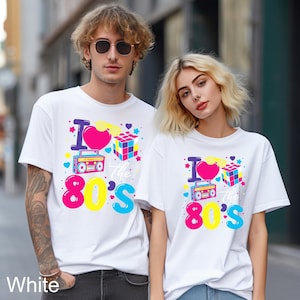 May include: Two white t-shirts featuring a colorful graphic print that says I Love the 80's. The design includes a pink heart, a yellow pair of sunglasses, a multi-colored puzzle cube, and a blue and pink boombox with various small star shapes.