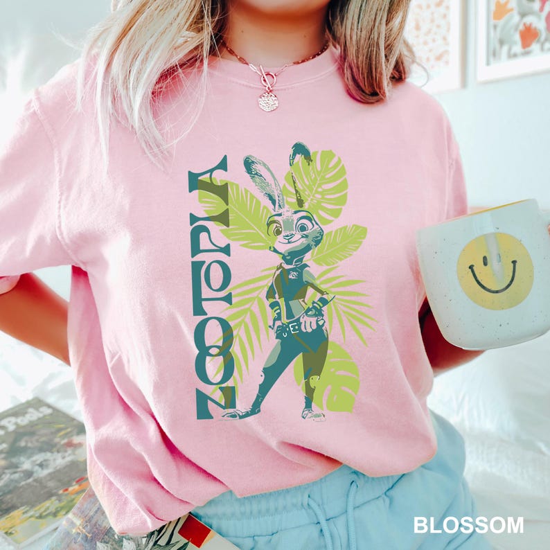 May include: Pink t-shirt featuring the word "Zootopia" in teal, with a cartoon image of Judy Hopps and tropical leaves. A white mug with a yellow smiley face is held in the hand. The word "BLOSSOM" is printed in white.