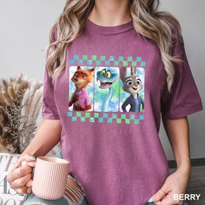 May include: Berry-colored t-shirt featuring three cartoon characters: a fox in a pink shirt, a snake, and a rabbit in a blue jacket. The characters are set against a colorful background and framed by a checkered pattern. The word "BERRY" is printed at the bottom.